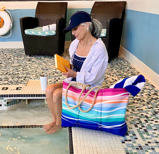 An older woman sits by an indoor pool with her feet in the water, holding a book, while the Sea Bags Sunset Surf Ogunquit Beach Tote—with its colorful design, sturdy rope handles, and waterproof pockets—rests beside her. by Sea Bags