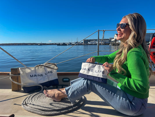 A woman in sunglasses and a green sweater sits barefoot on a boat holding the Sea Bags Maine Life Large Wristlet, with water, docks, and a clear blue sky in the background. by Sea Bags