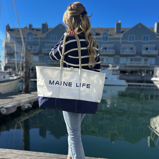 A woman with long, wavy hair holds the Maine Life Large Tote by Sea Bags near a marina, boats and waterfront buildings in the background on a sunny day—an effortless portrait that captures the spirit of Maine Life as curated by Erin Ovalle. by Sea Bags