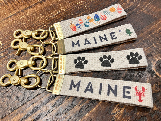 Four Rustic Marlin fabric bag charm keychains with brass hardware are arranged on a wooden surface. Each features a Maine-inspired design—mittens, pine trees, paw prints, or lobsters—and the word “Maine.” These Rustic Marlin Keychain Maine Lobsters make ideal Maine souvenirs. by Sea Bags