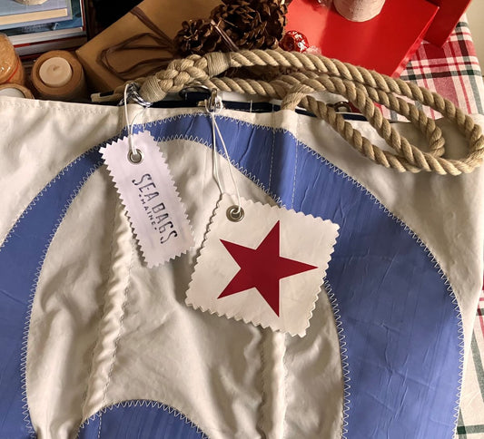 Elevate your holiday gifting with the Sea Bags Star Sail Cloth Gift Tag, featuring a unique red star and blue circle design on recycled sailcloth, accented by rope handles and two attached tags—one branded with Sea Bags and the other signifying its eco-friendly origins as a recycled sail. by Sea Bags