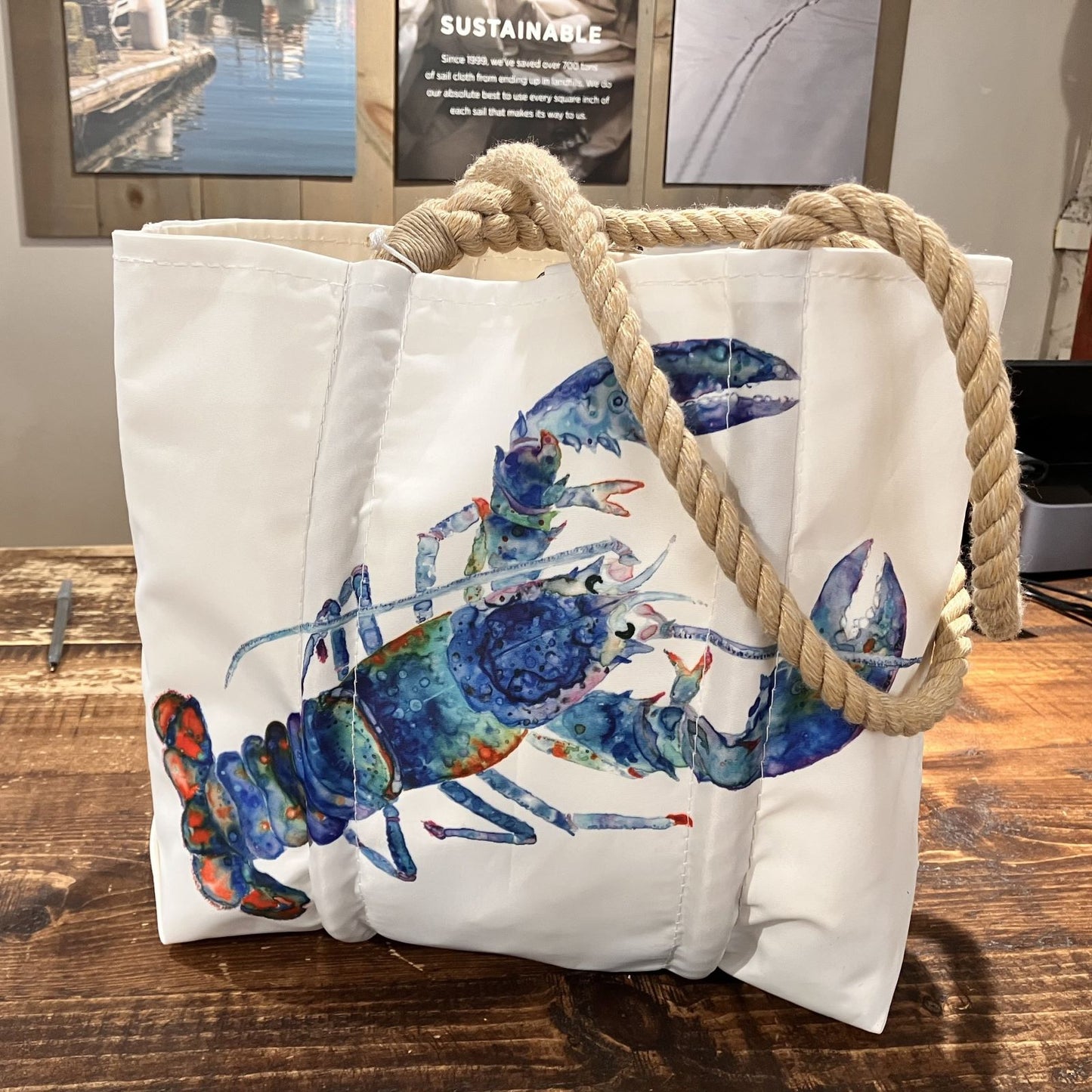 The Multicolor Lobster Handbag by Sea Bags, featuring a vibrant cotton candy lobster illustration by a Maine artist and sturdy rope handles, rests on a wooden table with framed pictures adorning the wall in the background. by Sea Bags