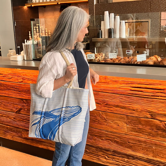A woman with gray hair stands at a bakery counter, carrying the Whale Watch Medium Tote by Sea Bags, handcrafted in Maine from recycled sailcloth. Pastries and coffee cups are visible behind the counter. by Sea Bags