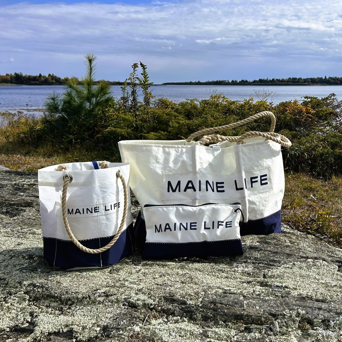 Three white and navy canvas bags labeled “MAINE LIFE” sit on a rock near a lake, with trees and a cloudy sky in the background—perfect for fans of the Maine Life TV show or anyone looking to carry their essentials in style, especially when paired with the Sea Bags Maine Life Large Wristlet. by Sea Bags