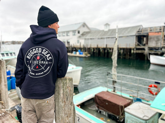 Wearing a Rugged Seas Navy Hoodie, a person stands by a dock, gazing at boats and a weathered building across the water on an overcast day. by Sea Bags