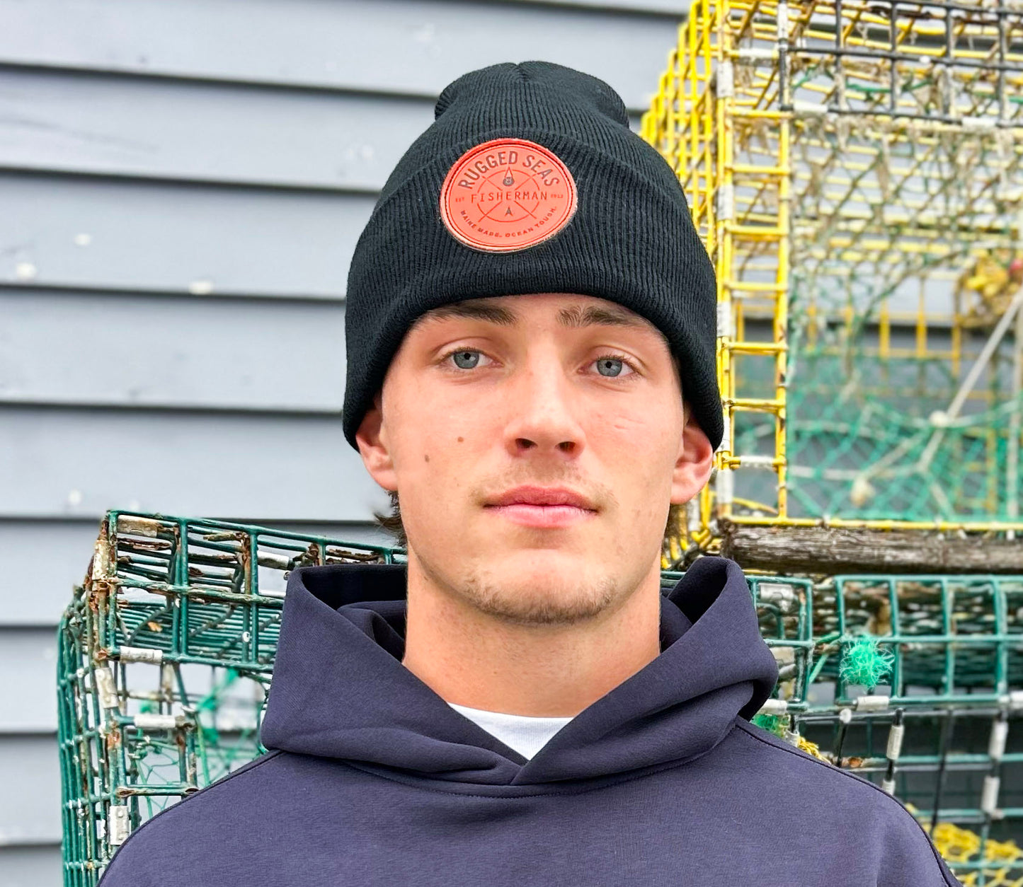 A young person wearing a Rugged Seas Watch Cap and navy hoodie stands before metal lobster traps and a gray wooden wall, making a nautical statement while looking directly at the camera. by Sea Bags
