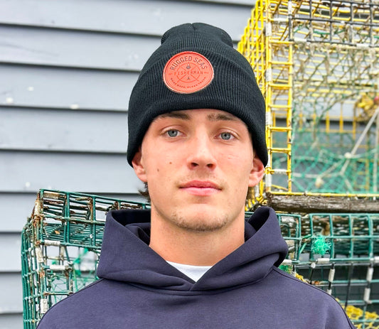 A young person wearing a Rugged Seas Watch Cap and navy hoodie stands before metal lobster traps and a gray wooden wall, making a nautical statement while looking directly at the camera. by Sea Bags