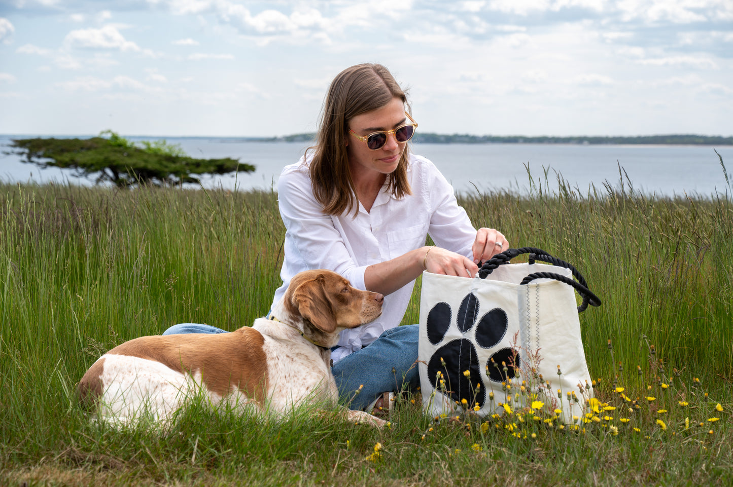 A woman in sunglasses sits on the grass by the water with her dog, reaching into her Sea Bags Paw Print Medium Tote with Clasp and Hanging Pocket—a white recycled sailcloth bag featuring a bold black paw print. Yellow flowers surround them. by Sea Bags
