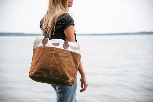 A woman stands by the water holding a Sea Bags Tan Chebeague Tote, crafted from recycled sail cloth, while wearing a black shirt and jeans. by Sea Bags