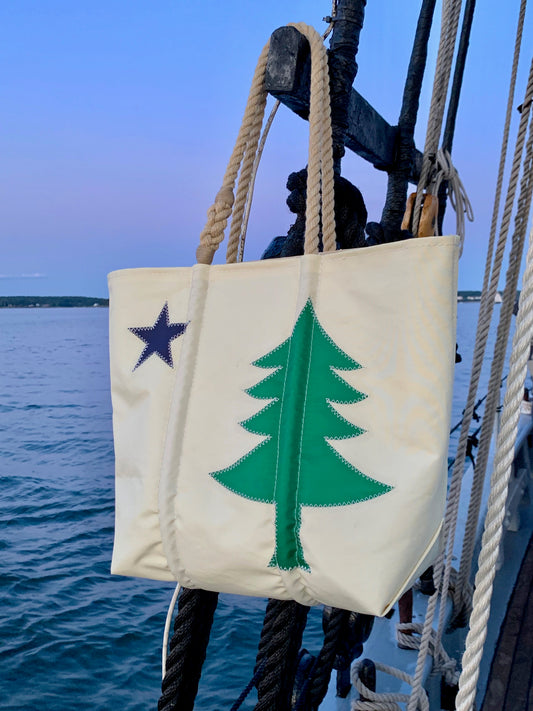 A Sea Bags Maine Bicentennial Medium Tote, crafted from recycled sail cloth with rope handles and adorned with a green pine tree and a blue star inspired by the Maine state flag, hangs on the railing of a boat at sunset. by Sea Bags