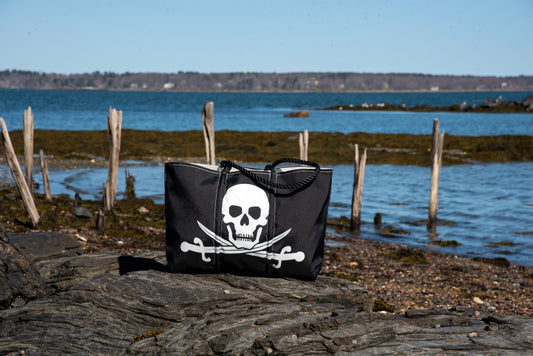 The Sea Bags Jolly Roger Tote with Clasp and Hanging Pocket, made from recycled sail cloth, showcases a bold white skull and crossed swords design on its black exterior. The tote is pictured resting on rocks by the shoreline, with the sea and a wooden fence in the background. by Sea Bags
