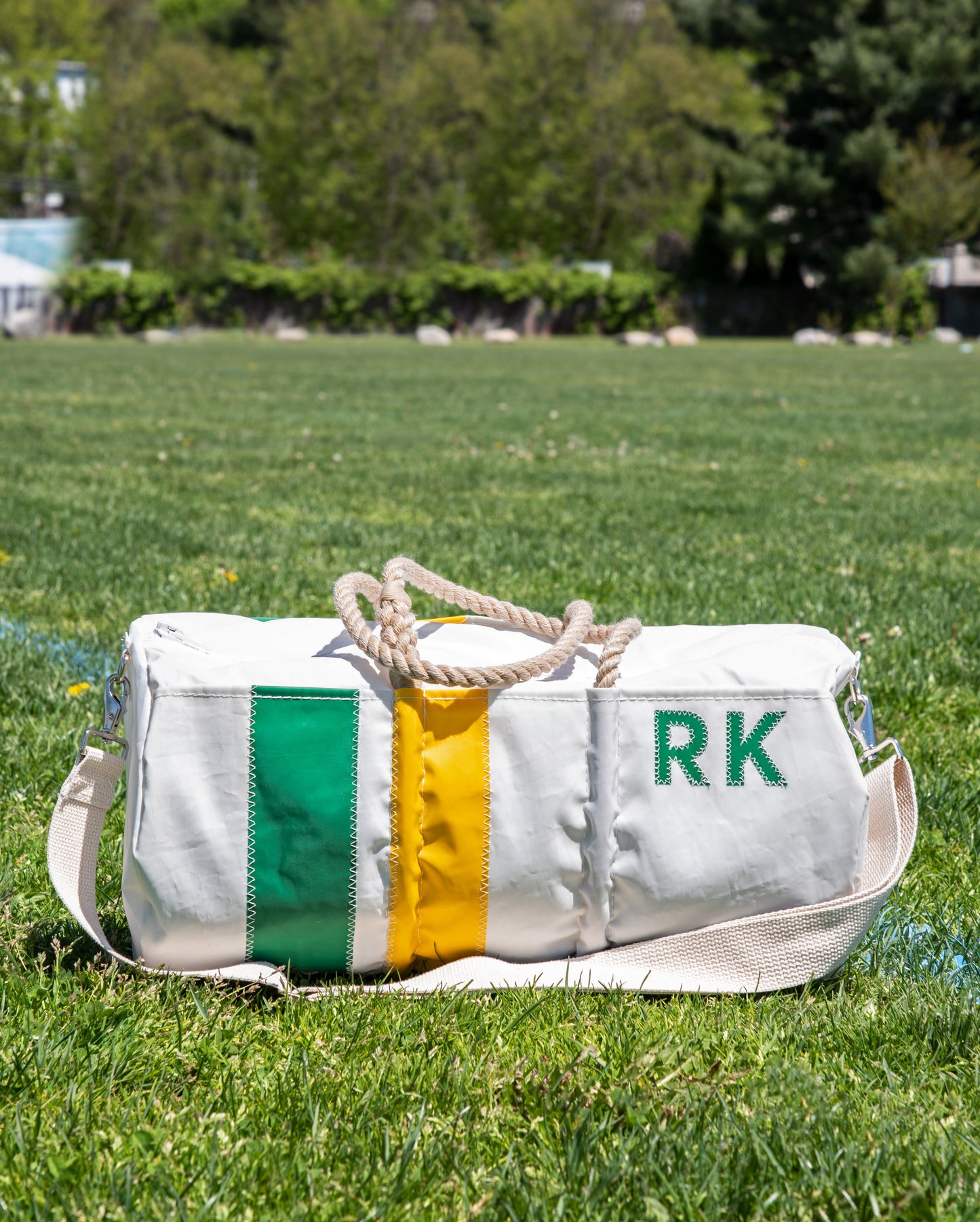 A Sea Bags Custom Monogram Vertical Stripe Duffel, made from white recycled sail cloth with green and yellow stripes, rope handles, and the initials RK, sits on grass with trees in the background. by Sea Bags