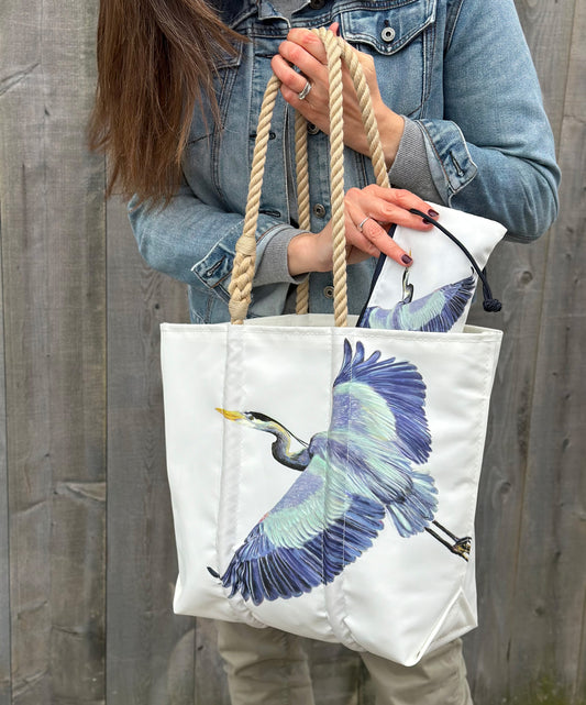 A person holds the Sea Bags Great Blue Heron Medium Tote, made from recycled sail cloth with rope handles and adorned with a blue heron illustration, along with a matching zip pouch—an ideal accessory for nature lovers—set against a wooden fence background. by Sea Bags
