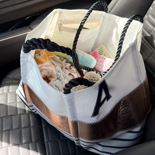 A Sea Bags Custom Monogram Rose Gold-on-Navy Mariner Stripe Large Tote, filled with towels, sunscreen, and a drink bottle, sits on a car seat. The bag features a prominent monogram on the side, adding a personalized touch and nautical chic style. by Sea Bags