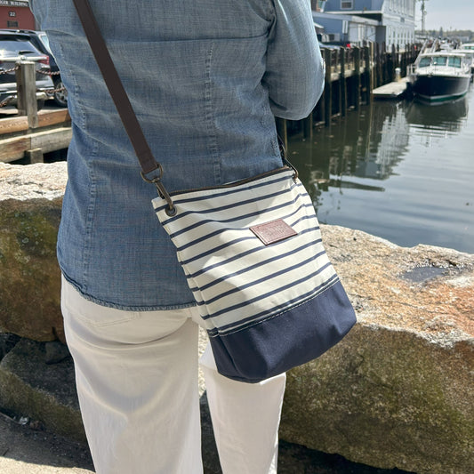 A person in a denim shirt and white pants stands by a waterfront, carrying the Sea Bags Navy Mariner Stripe Chebeague Crossbody Bag, crafted from recycled sailcloth. Boats and a stone barrier are visible in the background. by Sea Bags