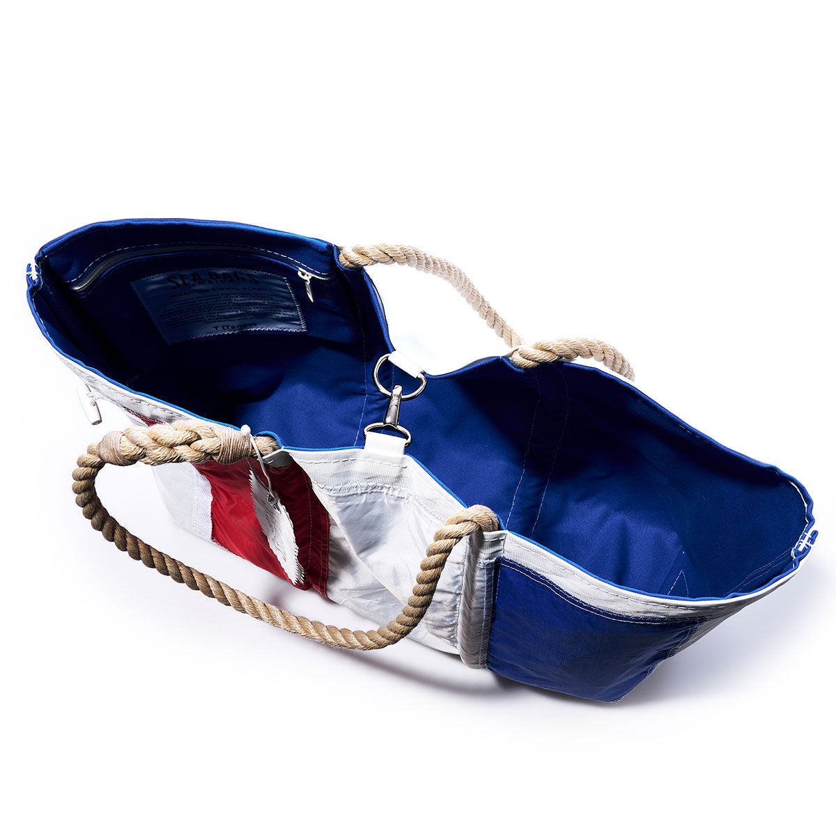 The Sea Bags Tango Signal Flag Large Tote, crafted from recycled sail cloth and inspired by maritime signal flags, features bold blue, white, and red panels, sturdy rope handles, and a blue interior lining. Shown from above with its top open, this bag blends nautical heritage with sustainable style. by Sea Bags