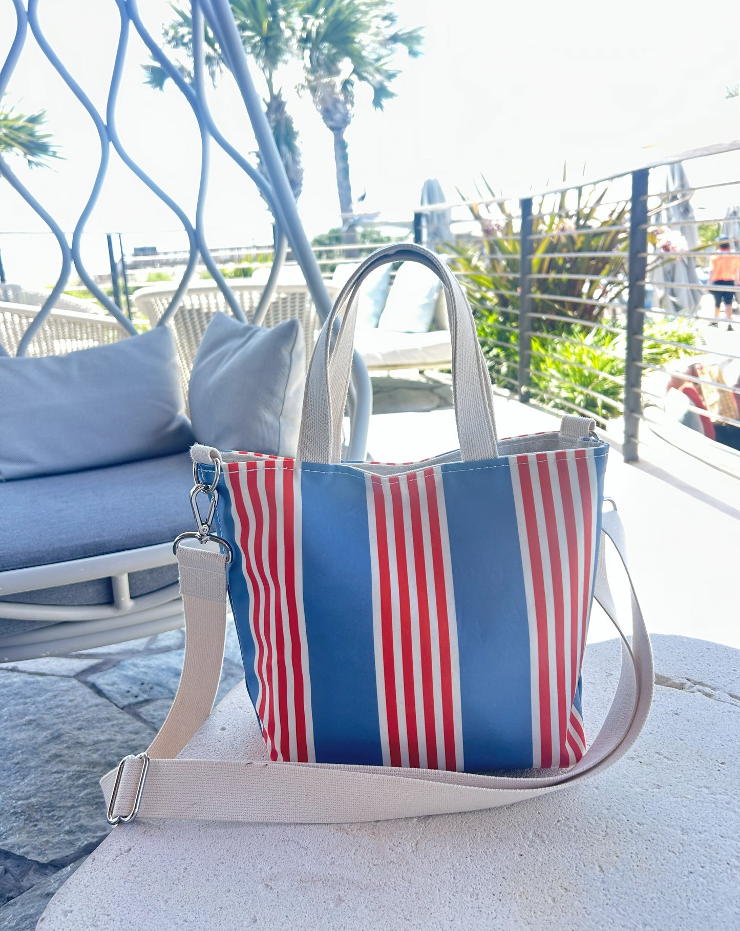 The Sea Bags French Stripe Crossbody Tote, featuring a red, white, and blue French stripe design with beige straps, rests on a round table outdoors near a lounge chair amidst lush greenery and palm trees. by Sea Bags