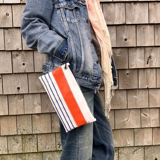 A person in a denim jacket and jeans stands in front of a shingled wooden wall, holding the Sea Bags Trapline Mariner Stripe Large Wristlet - Orange, a rectangular bag featuring blue and orange stripes crafted from recycled sail cloth. by Sea Bags