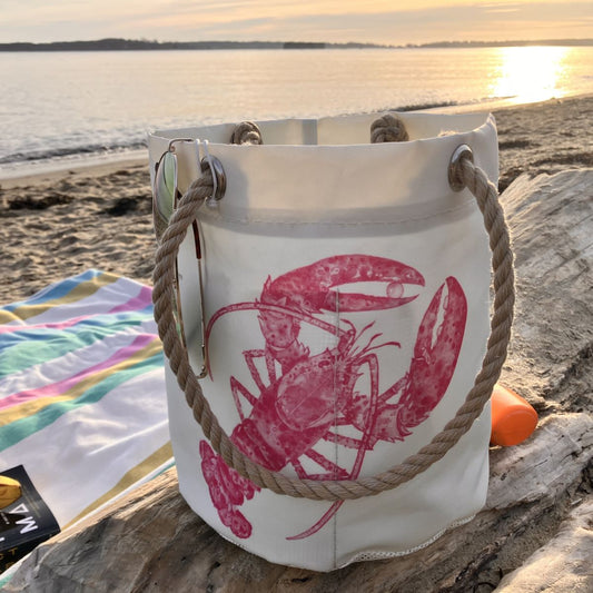 The Sea Bags Pink Lobster Beachcomber Bucket, made from recycled sail cloth and featuring pink lobster prints with sturdy rope handles, rests on a driftwood log beside a towel and sunscreen, as the sun sets over a sandy beach and calm water. by Sea Bags