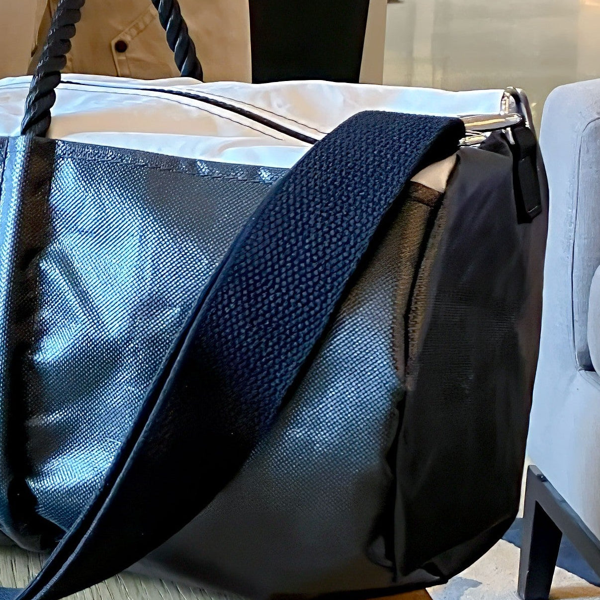 A close-up of the Sea Bags Black Dragon Duffel, featuring a silver and black textured design crafted from water-resistant recycled sail cloth, with a thick black strap, resting on a wooden surface next to a beige chair. by Sea Bags