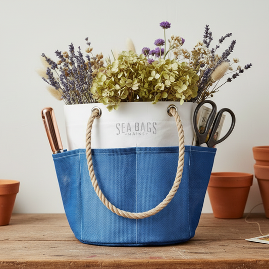 The Sea Bags Harbor Blue Gardeners Bucket Bag, featuring blue and white stripes and sturdy rope handles, holds dried flowers, scissors, and other gardening essentials. Its durable mesh sides keep contents visible inside, while empty terracotta pots are displayed in the background. by Sea Bags