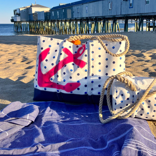 Sea Bags’ Fuchsia Anchor Summer Dots Ogunquit Beach Tote, featuring recycled sail cloth, sturdy rope handles, and a vibrant pink anchor motif with playful polka dots, rests on the sand near a pier—ocean waves and rustic wooden buildings forming the perfect coastal backdrop. by Sea Bags