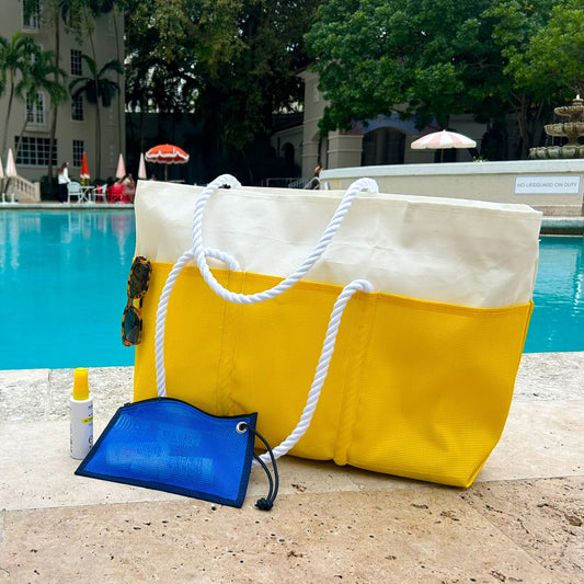 A Goldenrod Beachcomber Tote from Sea Bags, featuring sturdy rope handles and a yellow-and-white design, sits poolside alongside sunglasses, sunscreen, and a blue mesh pouch, with umbrellas, lounge chairs, and trees visible in the background. by Sea Bags