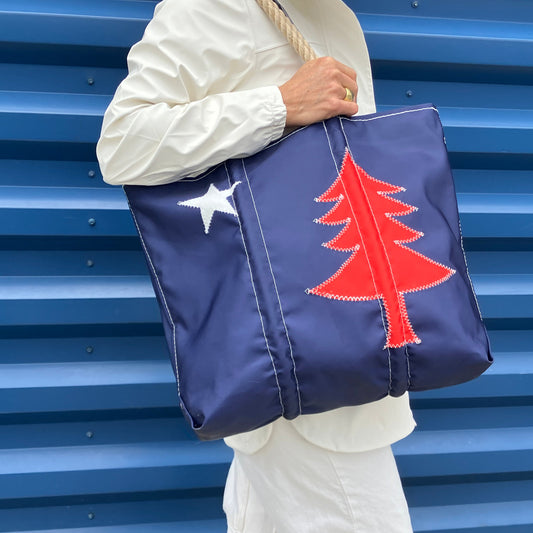 A person in a white outfit holds the Sea Bags 250th Patriotic Pine Medium Tote—crafted from recycled sailcloth and featuring a white star and red pine tree—while standing against a blue, ridged wall. by Sea Bags