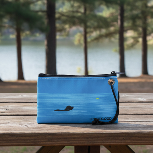 The Sea Bags Life is Good Dog Days Wristlet, featuring a blue design with a black dog graphic, rests on a wooden picnic table by a lakeside surrounded by trees—making it the perfect eco-friendly handbag or wristlet for any adventure. by Sea Bags