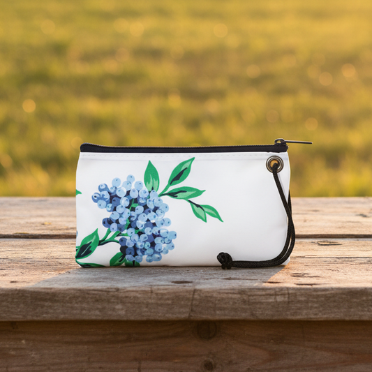 Sea Bags Blueberry Print Wristlet, crafted from recycled sail cloth and adorned with a blue and green floral design inspired by Maine blueberries, features a black zipper and wrist strap. Photographed outdoors on a wooden surface with blurred grass in the background. by Sea Bags