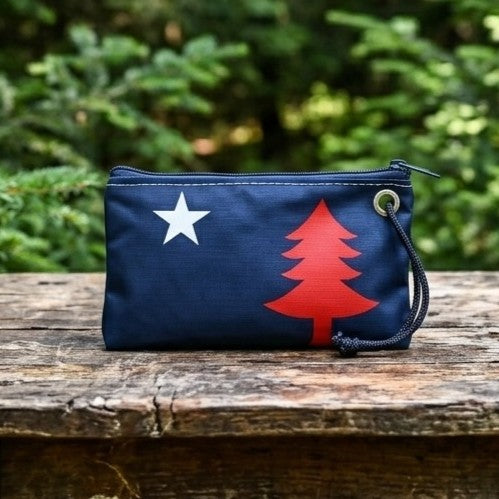 The 250th Patriotic Pine Wristlet by Sea Bags, crafted from recycled sailcloth in navy blue and adorned with a white star and red pine tree design, sits on a wooden surface with greenery in the background—a perfect patriotic bag or Maine wristlet for any occasion. by Sea Bags
