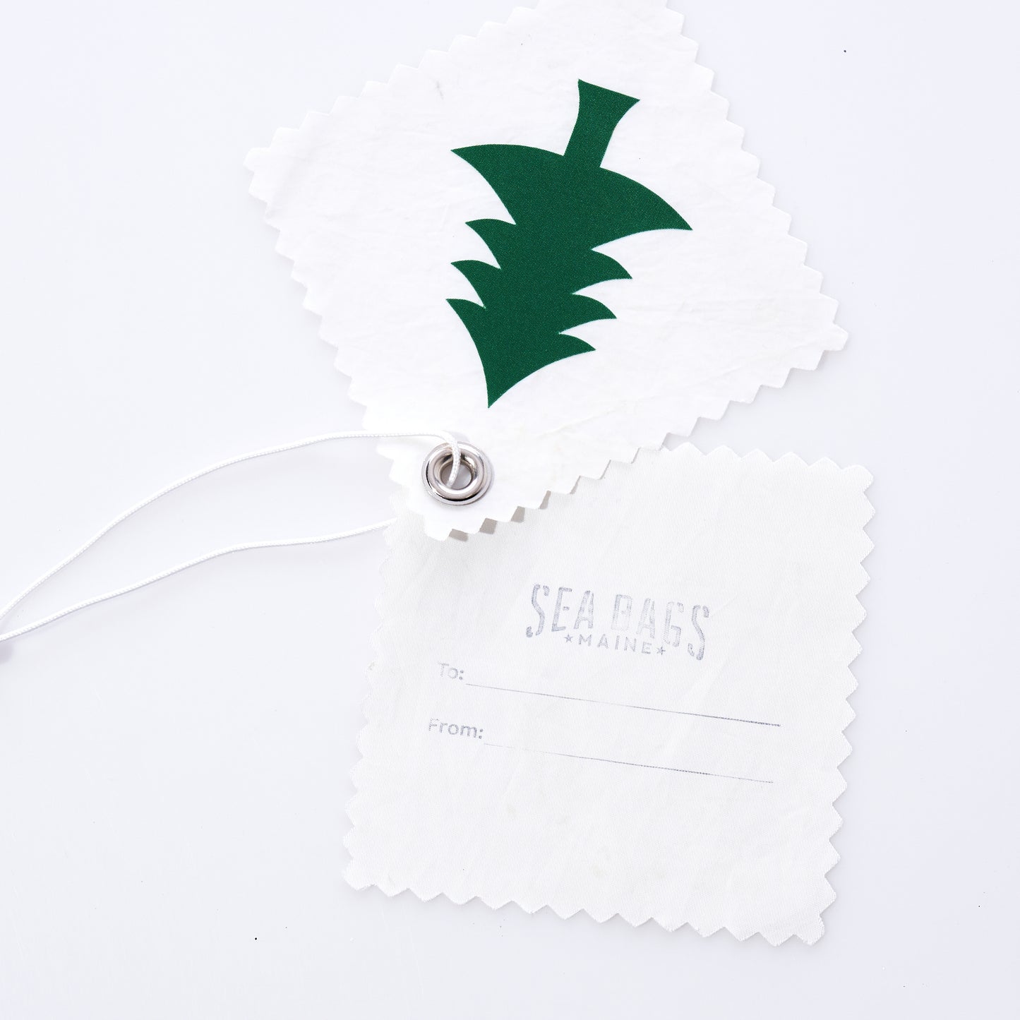 Two white, textured Pine Tree Sail Cloth Gift Tags by Sea Bags—made from recycled sails and attached by string—include one with a green pine tree design perfect for gifting, and the other featuring faint Sea Bags Maine text with To and From lines, for a sustainable touch. by Sea Bags