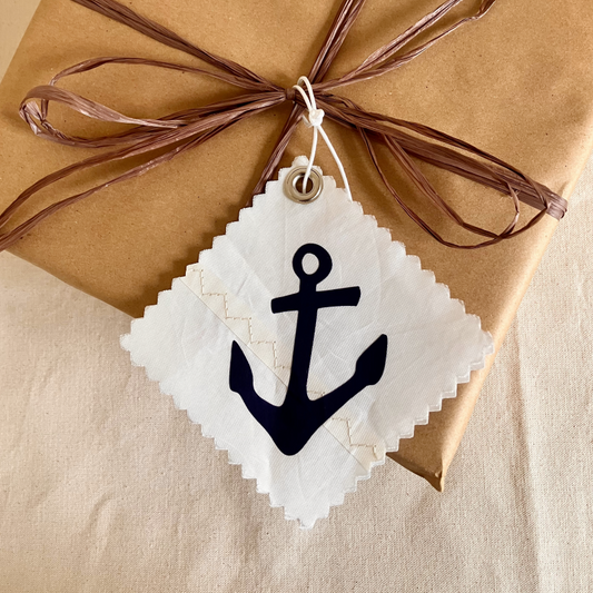 A brown gift wrapped with twine features a Sea Bags Navy Anchor Sail Cloth Gift Tag—a white square recycled sail tag displaying a black anchor symbol. by Sea Bags