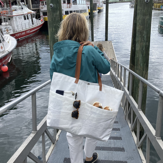A person wearing a blue jacket carries the Sea Bags Large Market Tote—crafted from white recycled sail with leather handles—down a dock ramp toward boats; the spacious tote holds sunglasses, cans, and a phone. by Sea Bags