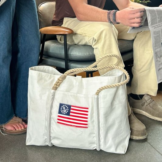 The Sea Bags U.S. Yacht Ensign Wharf Tote, made from recycled sail cloth and featuring sturdy rope handles along with an early U.S. flag design, rests on the floor near two people—one holding a newspaper—embodying sustainable craftsmanship and timeless nautical style. by Sea Bags