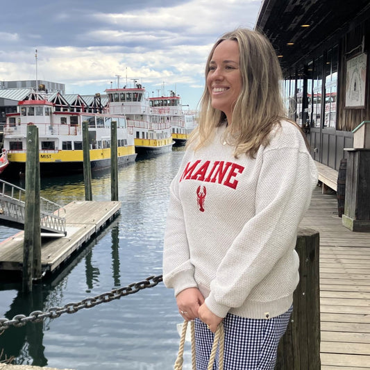Wearing a Soft As A Grape Popcorn Fleece Pullover in Oatmeal featuring the Maine Lobster design, a person stands by a dock with yellow and white ferries in the background on a cloudy day. by Sea Bags