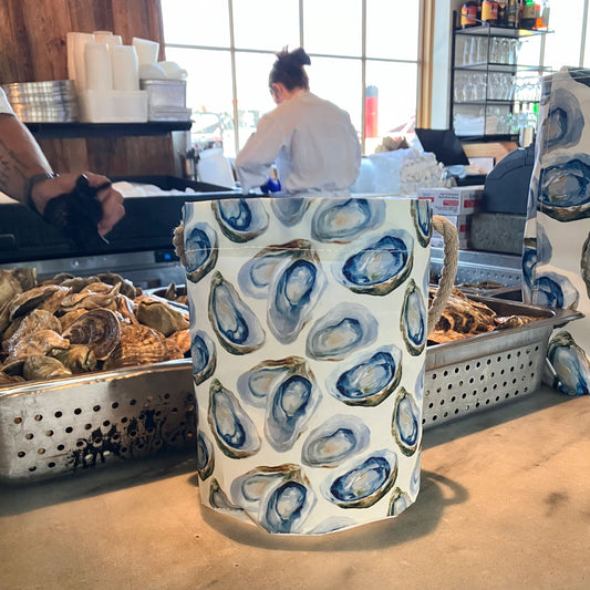 A ceramic pitcher adorned with blue and gray oyster shell patterns sits on a counter near fresh oysters in a restaurant kitchen, evoking the watercolor oyster design featured on the Watercolor Oyster Bucket by Sea Bags, which is crafted from recycled sail cloth.