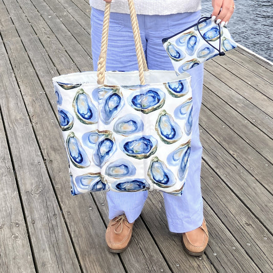 A person stands on a wooden dock holding the Sea Bags Watercolor Oyster Medium Tote and a matching pouch, both featuring blue oyster shell patterns. by Sea Bags