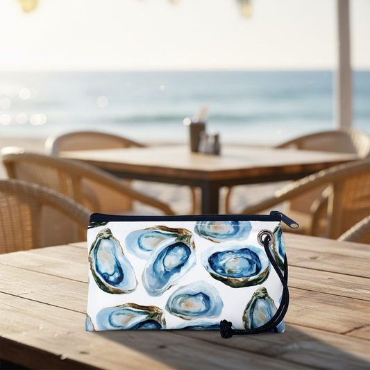 A Sea Bags Watercolor Oyster Wristlet rests on a wooden table at an outdoor beachside café, with empty chairs and the ocean in the background. by Sea Bags