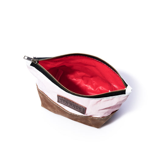 The Tan Chebeague Cosmetic Bag by Sea Bags is a small zippered bag made from recycled sailcloth, featuring a white and tan exterior, red interior lining, and a “SEA BAGS” label on the front; it is shown open against a white background. by Sea Bags