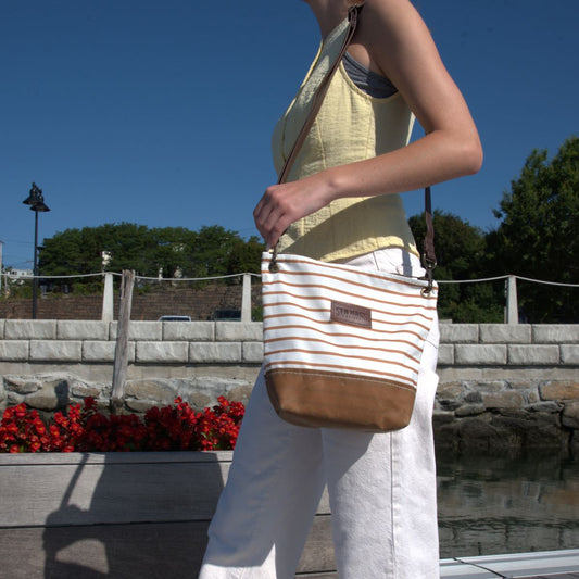 A person wearing a yellow top and white pants carries the Sea Bags Tan Chebeague Stripe Crossbody, featuring a leather shoulder strap and crafted from recycled sailcloth, outdoors on a sunny day.