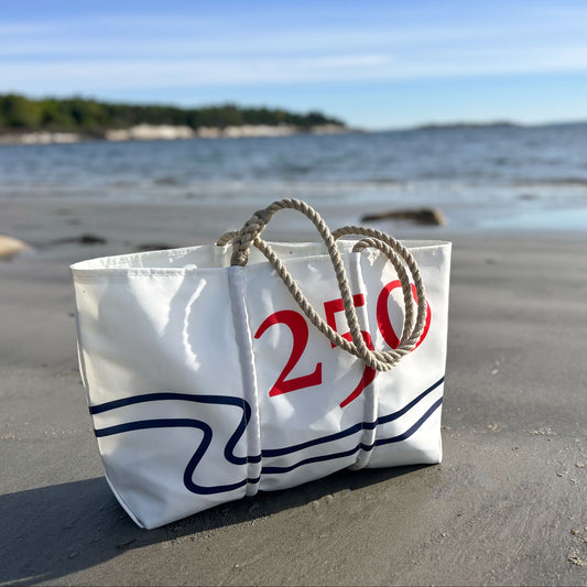 The Sea Bags 250th Patriotic Large Tote, crafted from recycled sail cloth and adorned with 250 in bold red, features sturdy rope handles and sits on a sandy beach with ocean and trees in the background. This handcrafted tote proudly showcases its Maine-made heritage. by Sea Bags