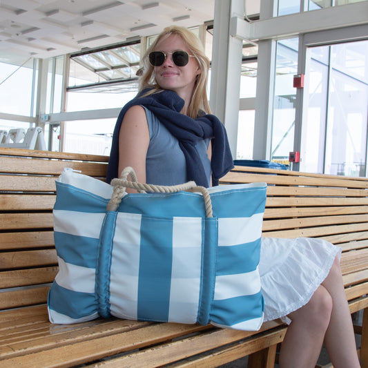 A woman wearing sunglasses and a navy sweater sits on a wooden bench inside a bright, modern building, holding the Sea Bags Denim Blue Trolley Sleeve Travel Pier Tote made from recycled sailcloth with rope handles.