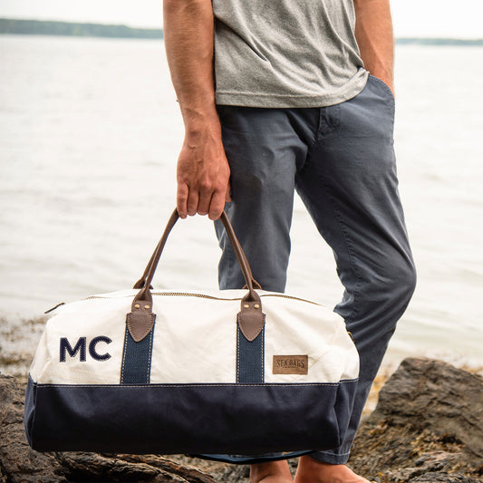 A person stands on rocks by the water, wearing a grey shirt and blue pants, holding the Sea Bags Custom Chebeague Duffel with brown handles and an MC monogram on the side. by Sea Bags