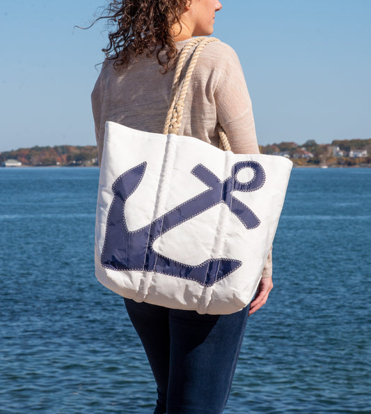 A person stands by the water, carrying the Sea Bags Navy Anchor Medium Tote with Hemp Rope—a white recycled-sail bag featuring a bold navy blue anchor design and sturdy hemp rope handles. by Sea Bags