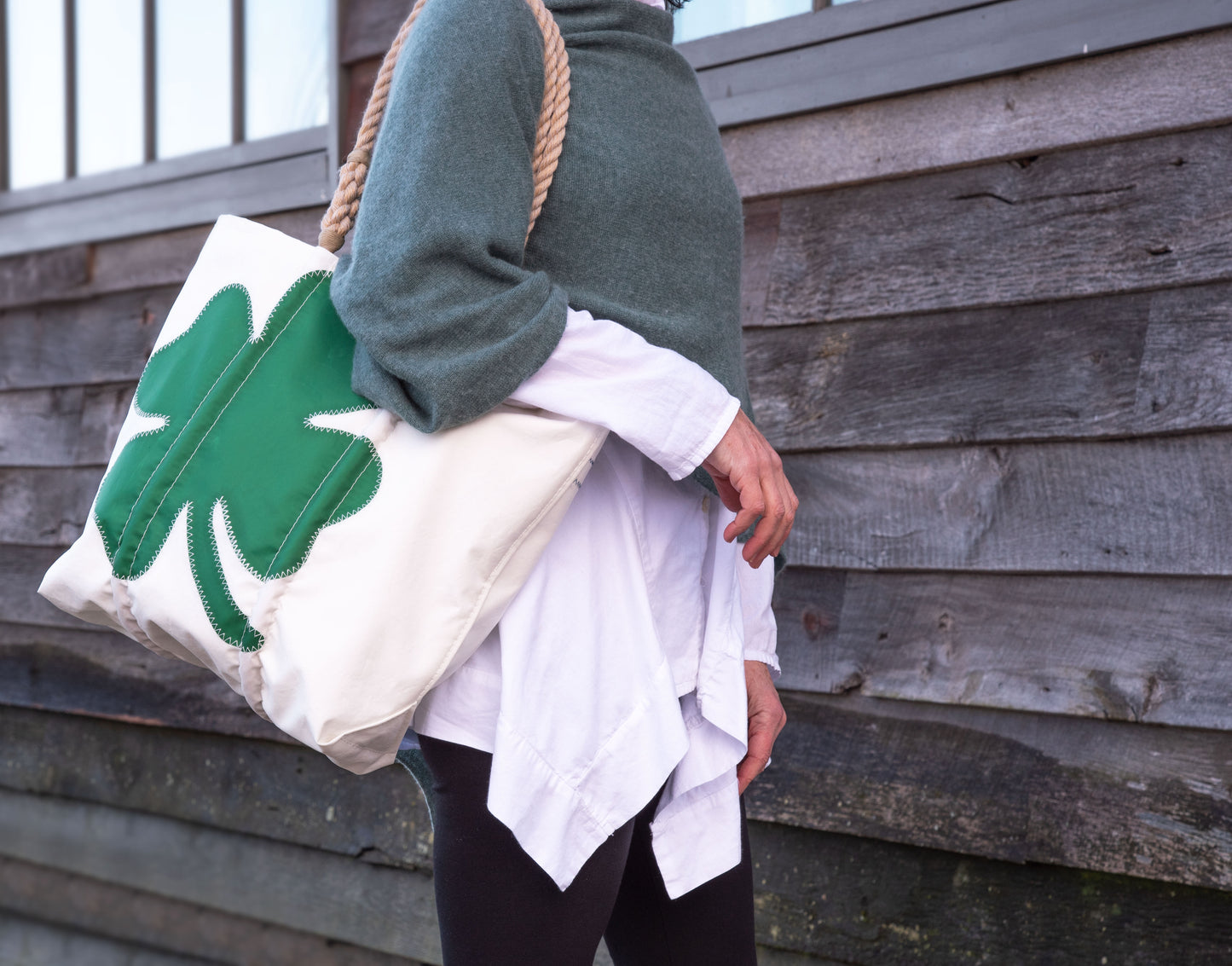 A person in a green sweater and white shirt carries the Sea Bags Four Leaf Clover Medium Tote—a spacious beige recycled sail cloth bag featuring a green four-leaf clover design, ideal for St. Patrick’s Day—while standing in front of a wooden wall. by Sea Bags