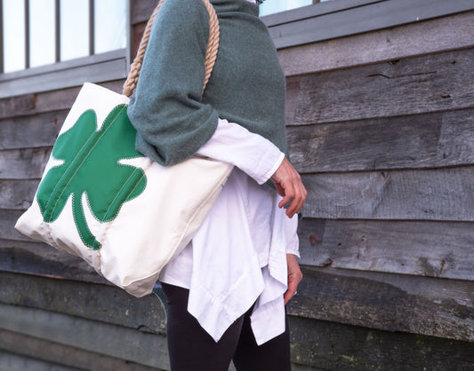 A person in a green sweater and white shirt carries the Sea Bags Four Leaf Clover Medium Tote—a spacious beige recycled sail cloth bag featuring a green four-leaf clover design, ideal for St. Patrick’s Day—while standing in front of a wooden wall. by Sea Bags