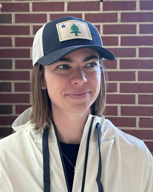 A person wearing a white jacket and the true Maine Flag Trucker Hat, which features a patch with a tree and blue star representing the Maine state flag, stands in front of a brick wall.