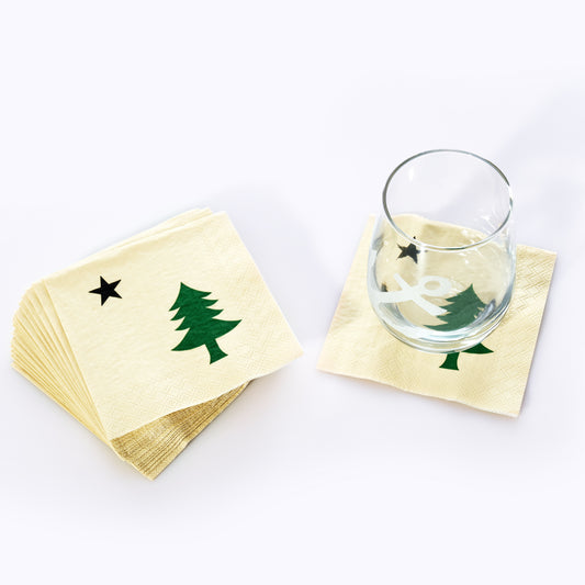 A glass rests on a Maine Bicentennial Cocktail Napkin by Boston International, decorated with a green Christmas tree and black star reminiscent of the pine-tree-and-star flag, while a stack of identical napkins sits nearby. by Sea Bags