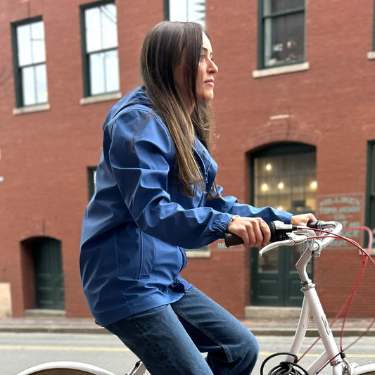 A woman wearing the Batela USA Inc Harborside Waterproof Raincoat in High Tide and jeans rides a white bicycle along a city street lined with red brick buildings. by Sea Bags